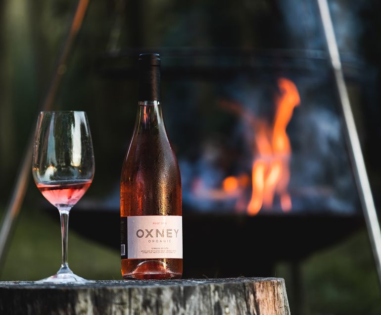 A bottle of Single Estate Rosé and a glass atop a stump of wood in front of a fire pit at Oxney Organic Estate